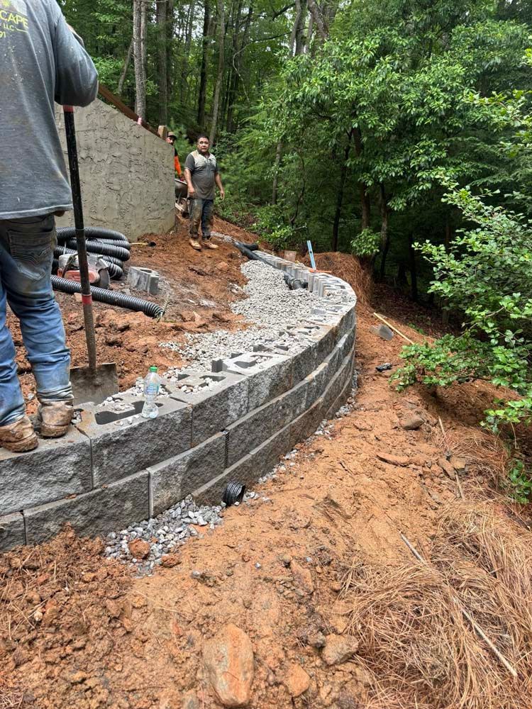 Curved gray segmental block retaining wall under construction with gravel backfill and drainage pipe on a wooded slope