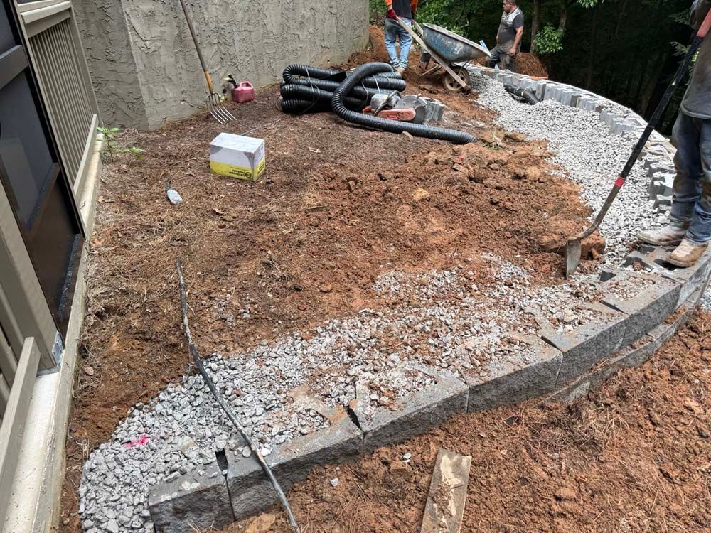 Segmental gray block retaining wall under construction with gravel backfill and drainage pipe beside a home