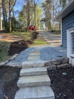 Natural stone step path with gray gravel walkway and boulder edging leading to a blue-gray home in a wooded yard