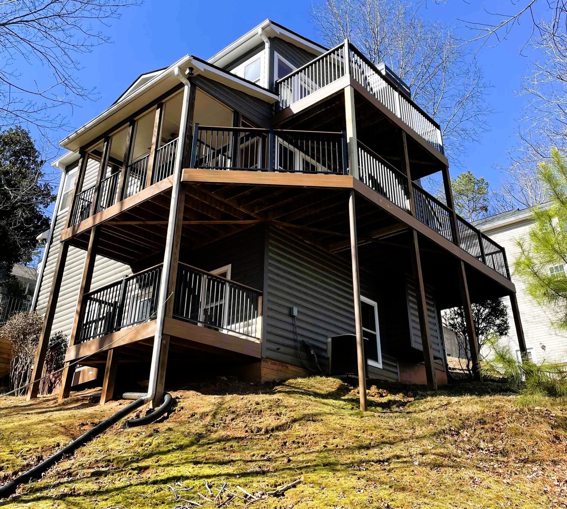 Multi-level stained wood deck with black metal balusters and screened porch on a hillside home under blue sky