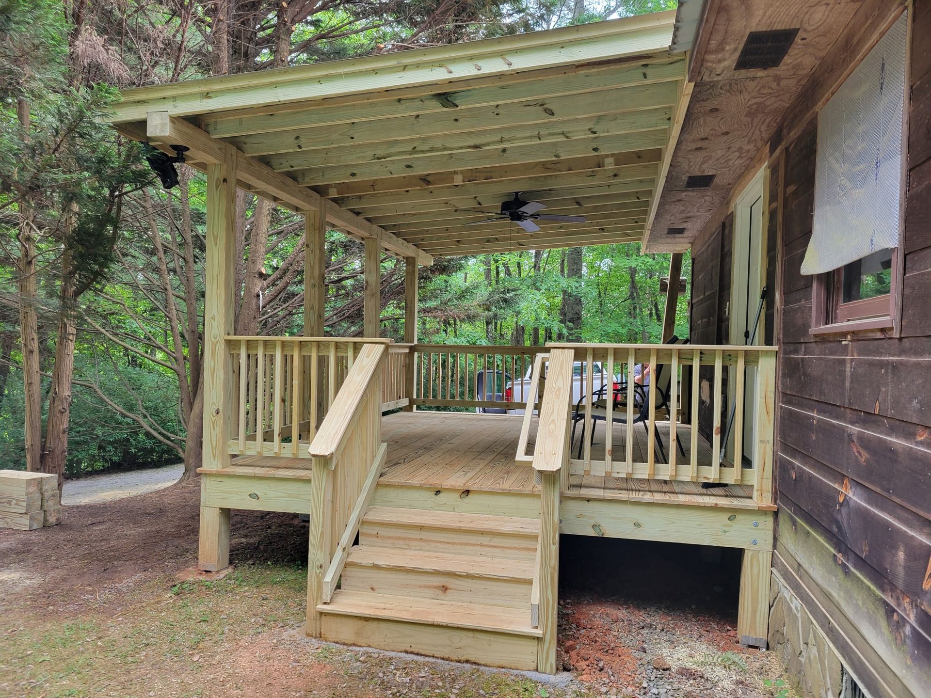 Covered pressure-treated wood deck with vertical baluster railings, steps, and ceiling fan attached to a wooded cabin
