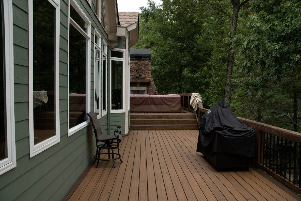 Brown composite deck with a covered hot tub on a raised platform and a stone outdoor fireplace beyond, beside a green-sided home