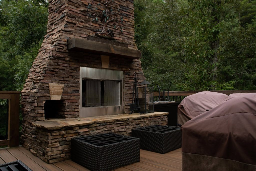 Stacked-stone outdoor fireplace with timber mantel built into a composite deck against dense woods