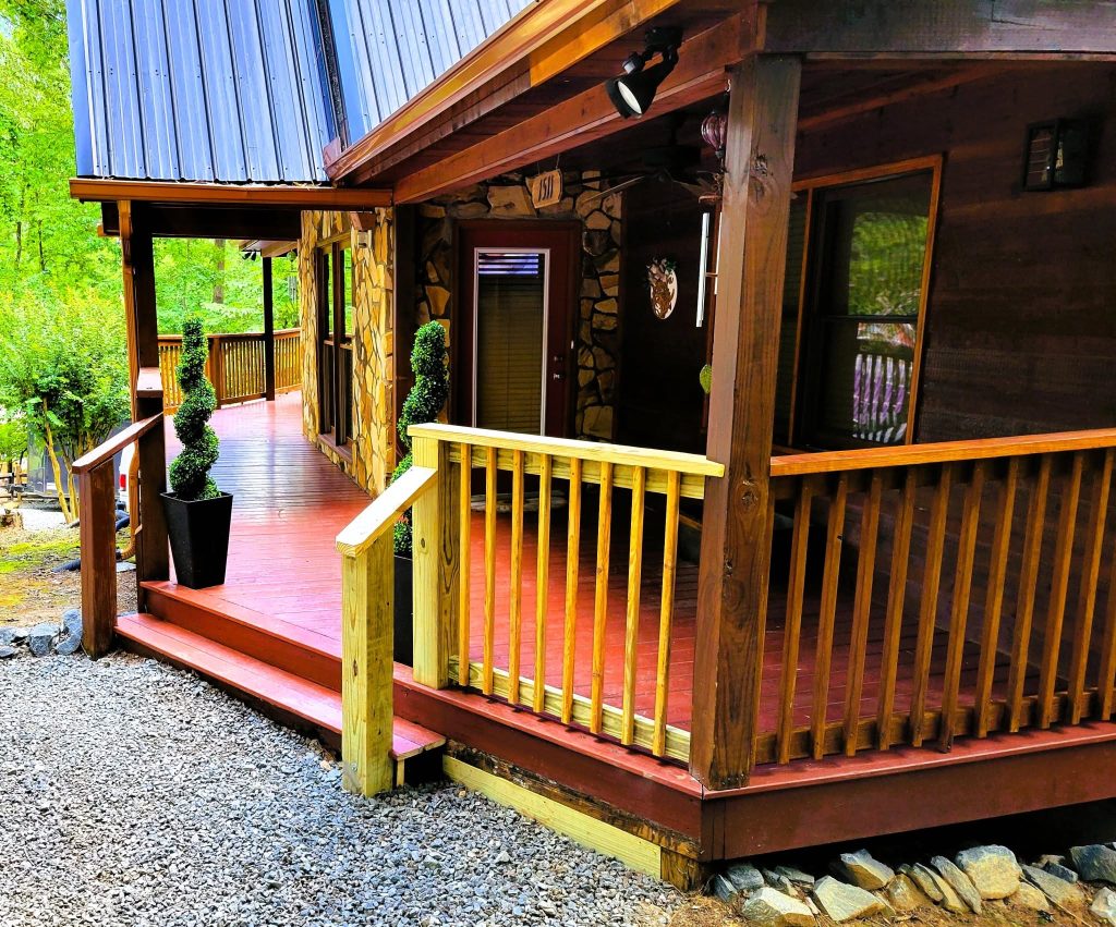 Wrap-around cabin porch with red-stained decking, stone column bases and a new pressure-treated railing section under a metal roof