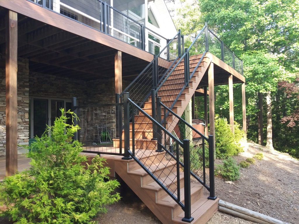 Composite stair run with black cable railing descending from an elevated deck past a dry-stack stone foundation into landscaped beds