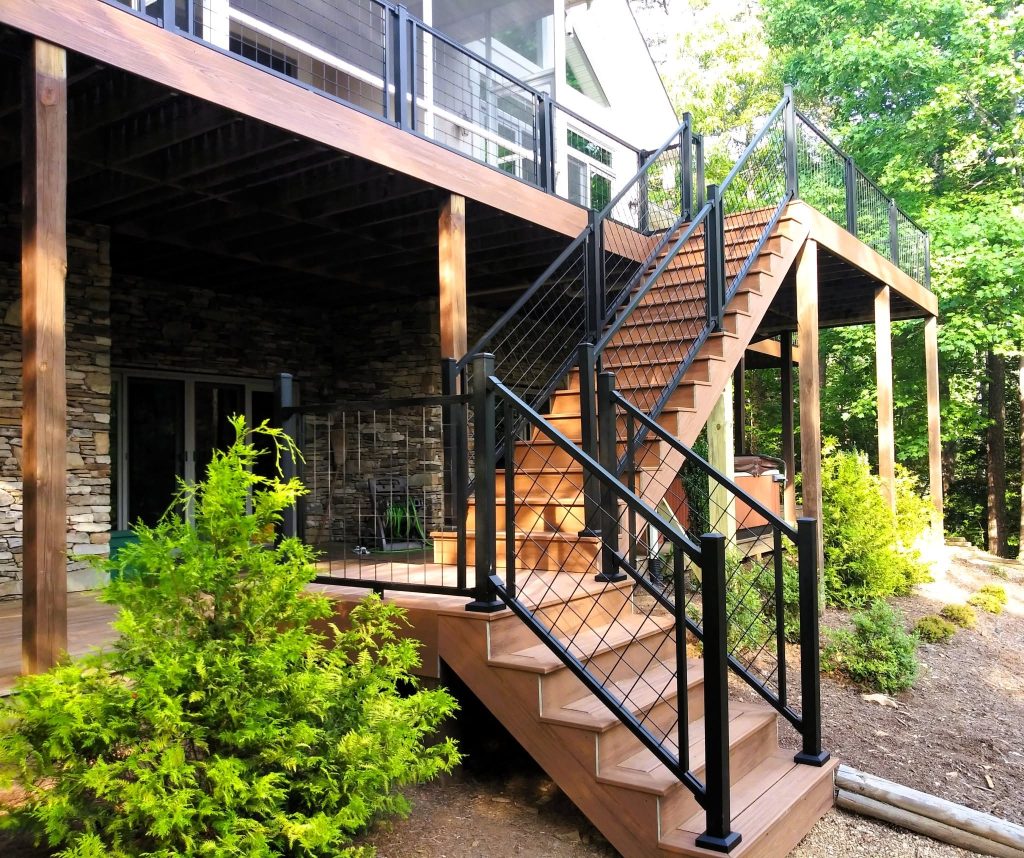 Wide composite stair run with black cable railing descending from an elevated deck past a dry-stack stone foundation