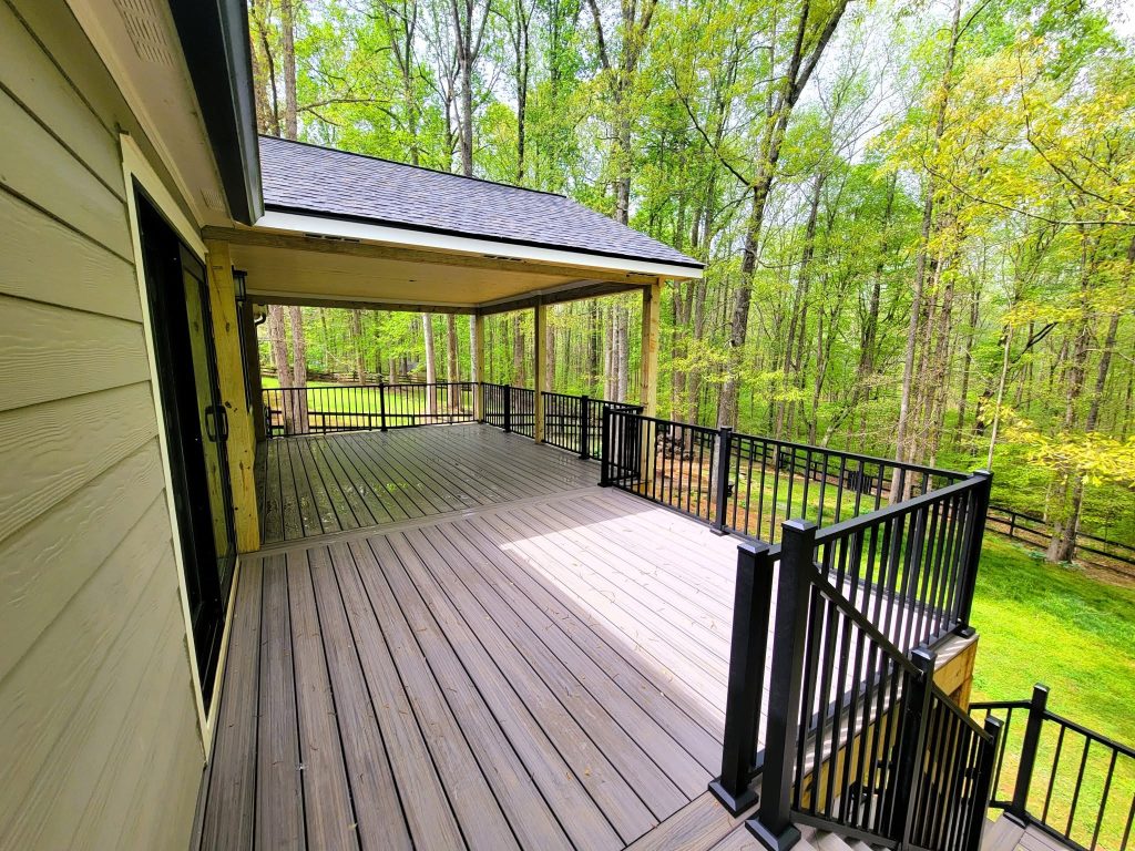 Covered deck with a hipped shingle roof, black aluminum railing and gray composite decking overlooking a fenced wooded lot