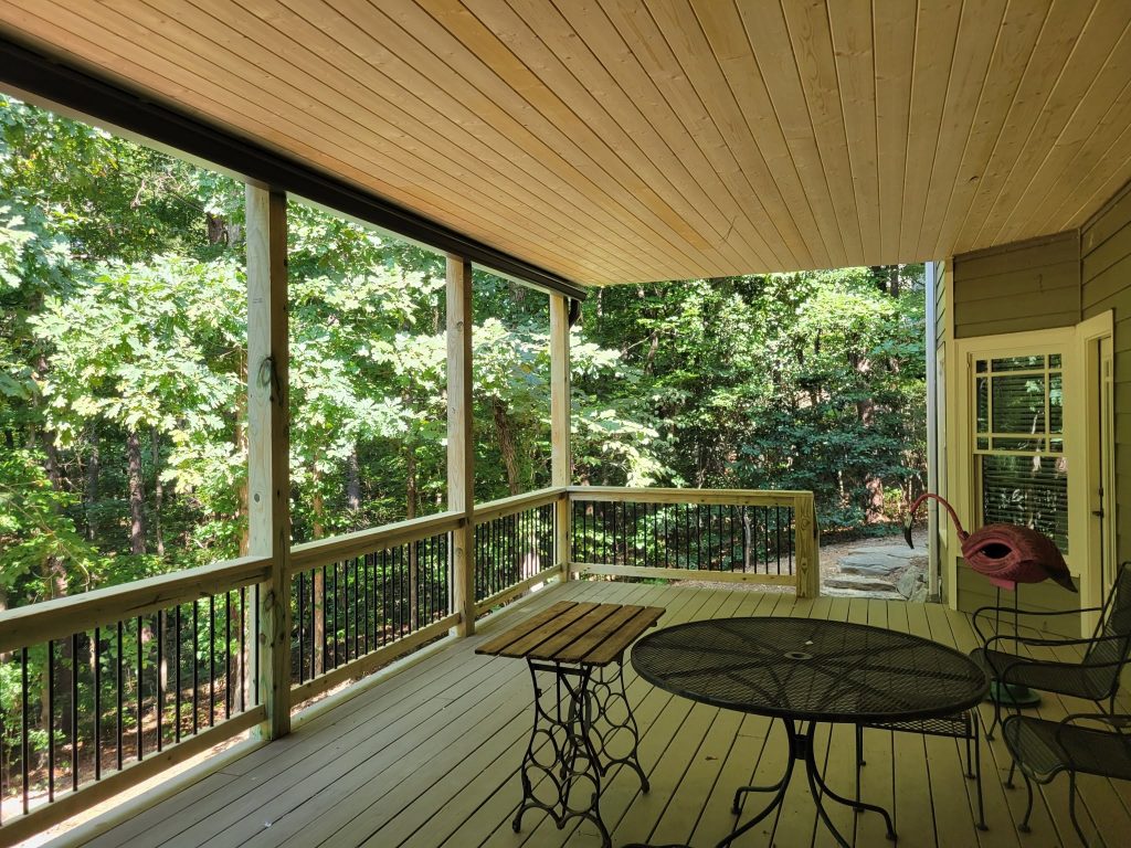 Covered porch with warm tongue-and-groove ceiling, black balusters and a metal bistro set in green woods