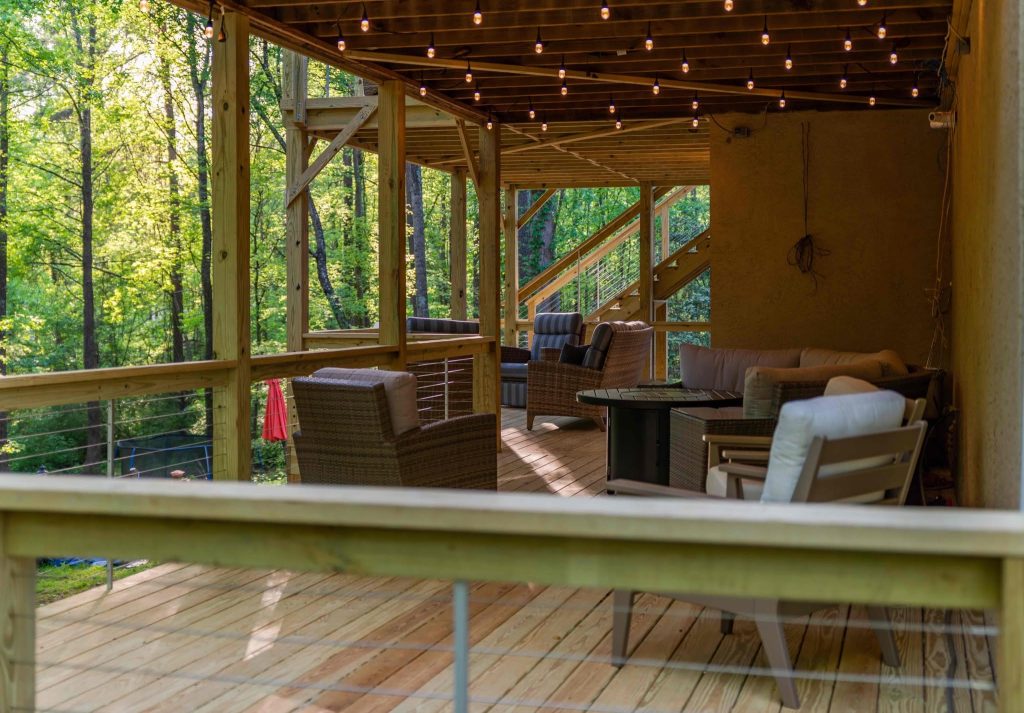Covered deck strung with festoon lights above cable railing and a wicker sectional in deep woods