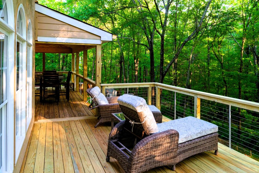 Cable-railed deck with two blue-cushioned wicker chaise lounges facing a wall of green forest