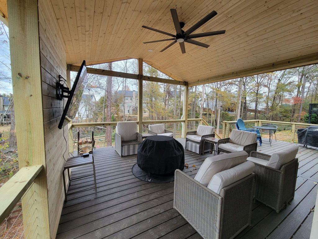 Covered deck with a pale pine cathedral ceiling, ceiling fan, mounted television and wicker seating around a covered fire pit