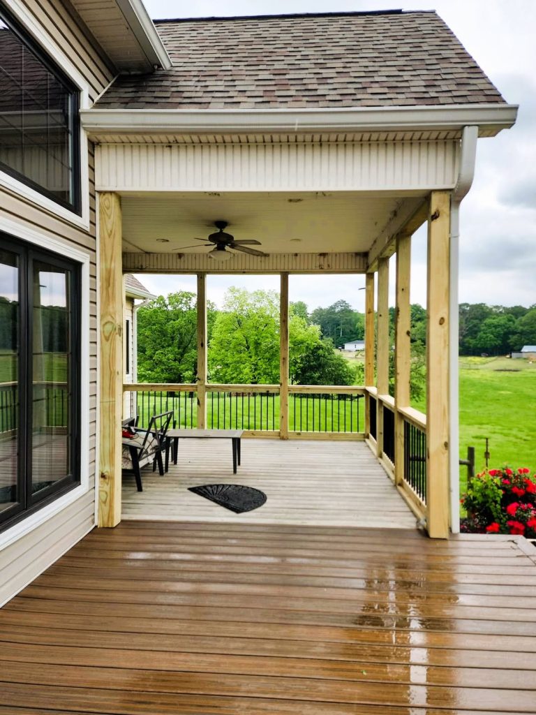 Covered porch with a beadboard ceiling and ceiling fan opening onto a wet brown composite deck above open pasture