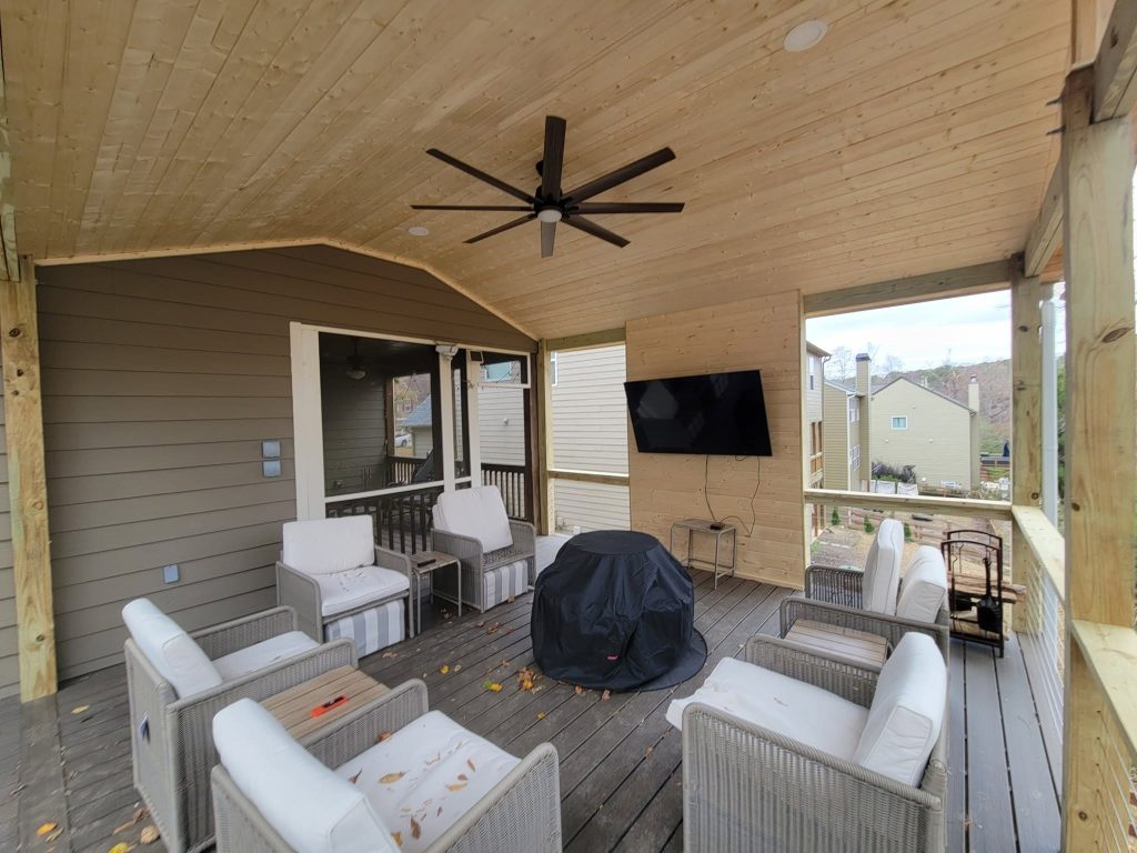 Covered porch with a curved tongue-and-groove pine ceiling, large ceiling fan, mounted television and wicker seating around a fire pit