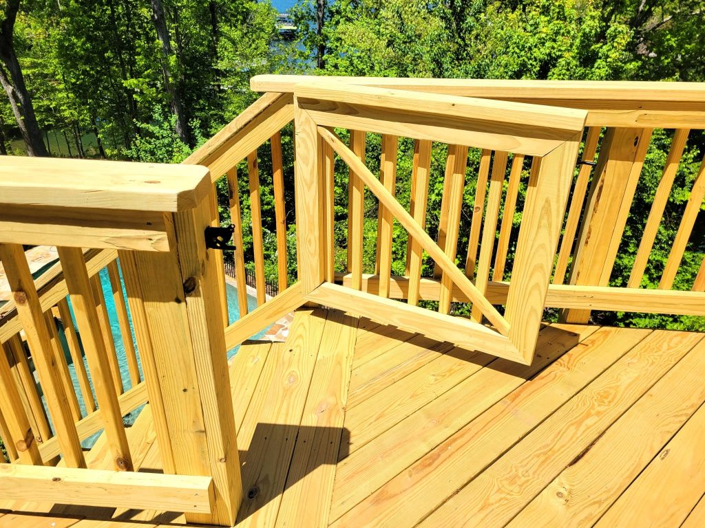 Open pressure-treated deck gate with diagonal bracing and latch hardware, above a pool with a lake beyond