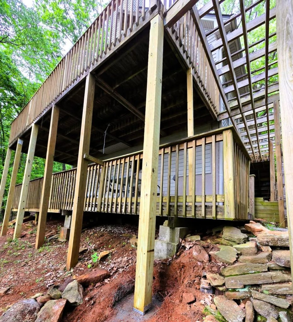 Upward view of a two-level deck substructure on tall pressure-treated posts with a pergola above and stone outcrop below