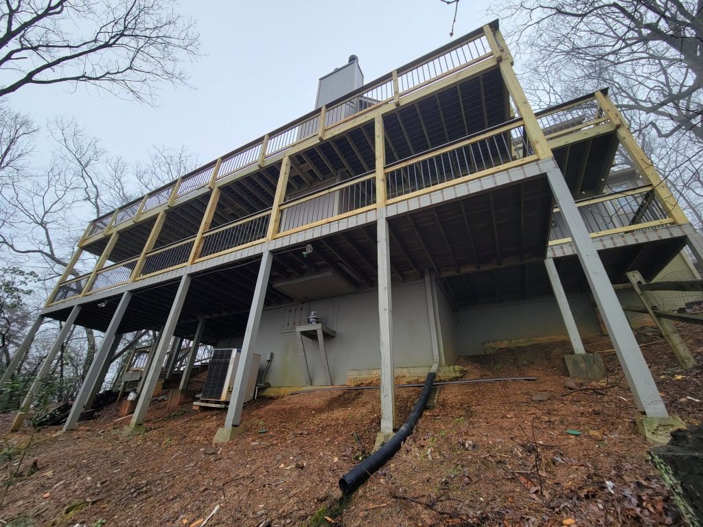 Upward view of a large elevated deck on tall posts with black baluster railing and full joist substructure on a wooded hillside