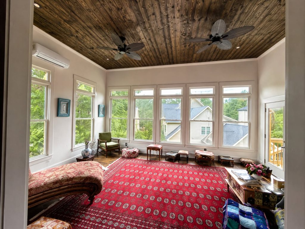 Enclosed sunroom with a dark stained tongue-and-groove ceiling, two ceiling fans, glazed walls and a red patterned rug