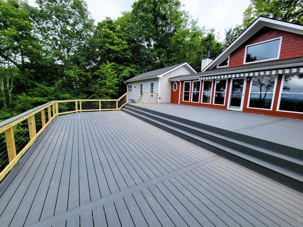 Wide gray composite deck with stepped tiers and cable railing wrapping a red shingled cabin among trees