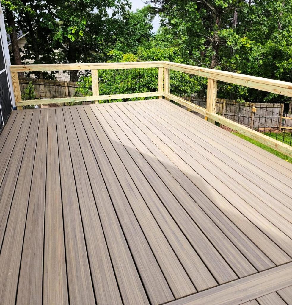 Gray-brown composite deck with picture-frame border boards and pressure-treated wire mesh railing beside a wooded fence line