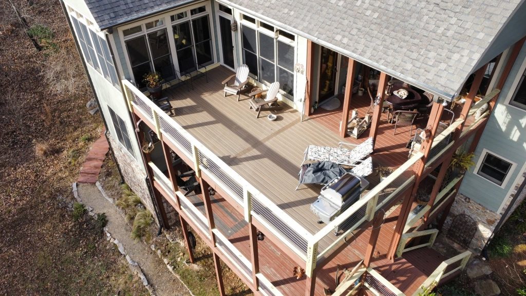 Aerial view of a dual-level gray composite deck with cable railing and Adirondack chairs on a wooded slope