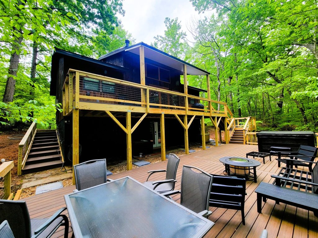 Multi-level deck with wire mesh railing, brown composite decking, a fire table and hot tub beside a dark cabin in dense woods