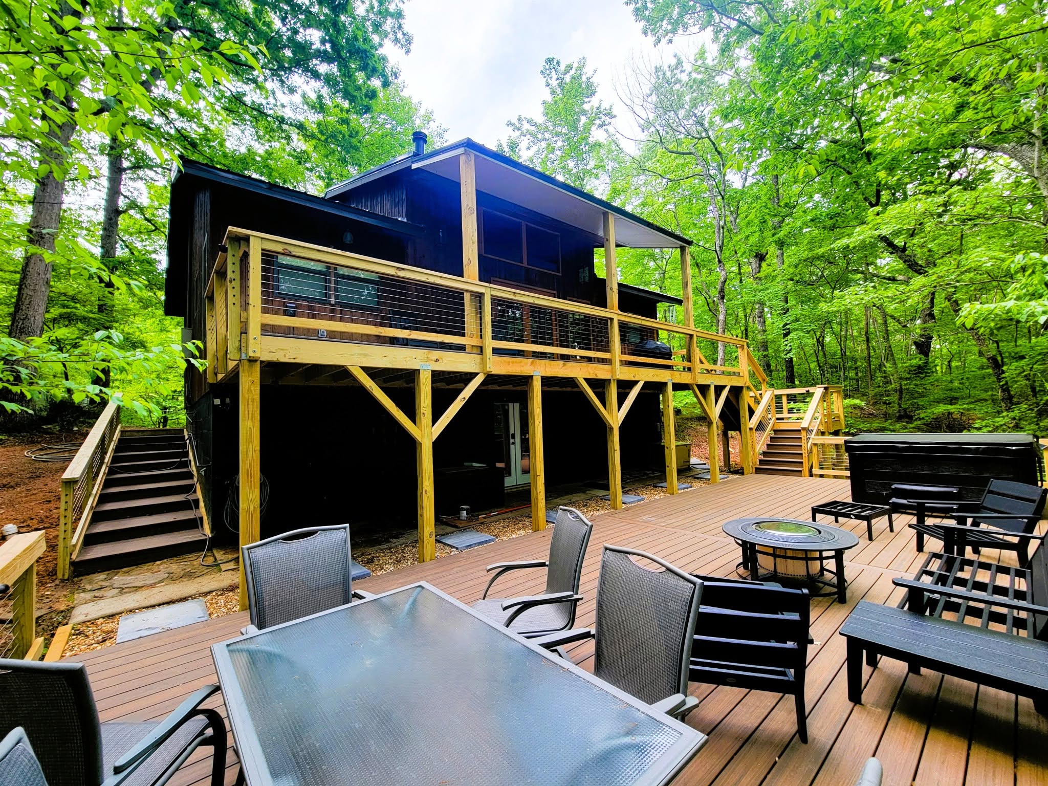 Multi-level deck with wire mesh railing, brown composite decking, a fire table and hot tub beside a dark cabin in dense woods