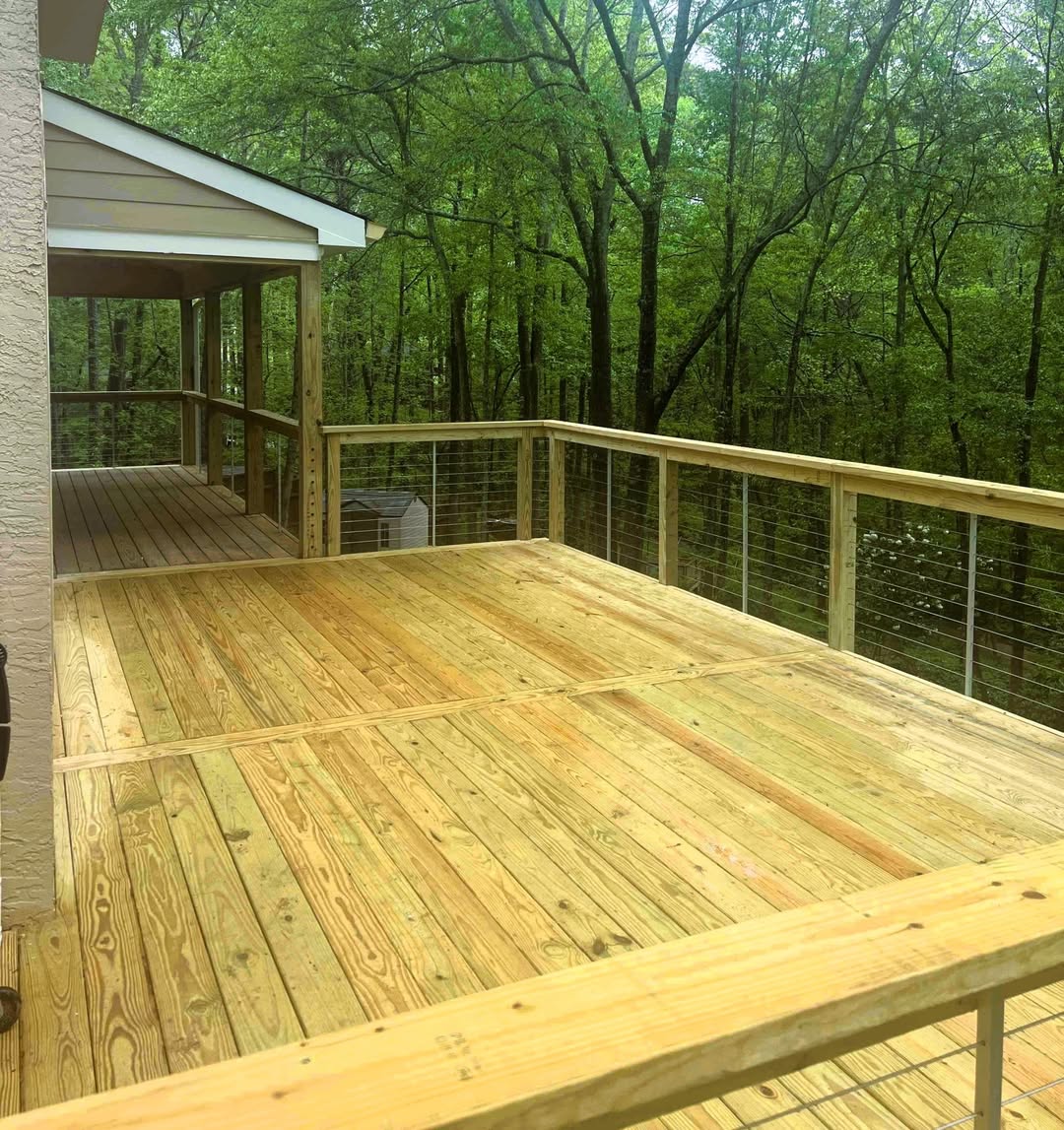 Newly built pressure-treated deck with cable railing extending from an existing screened porch into dense woods
