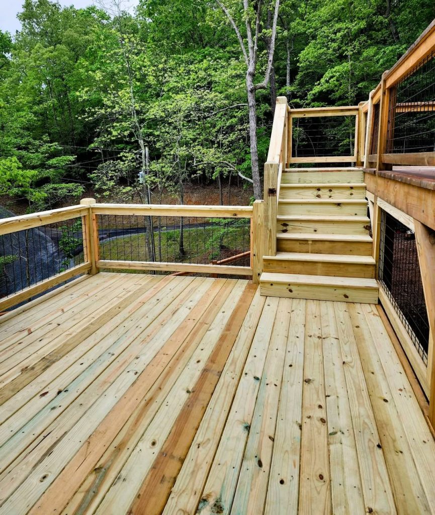 New pressure-treated deck with black wire mesh railing and a stair run rising to an upper level in dense woods
