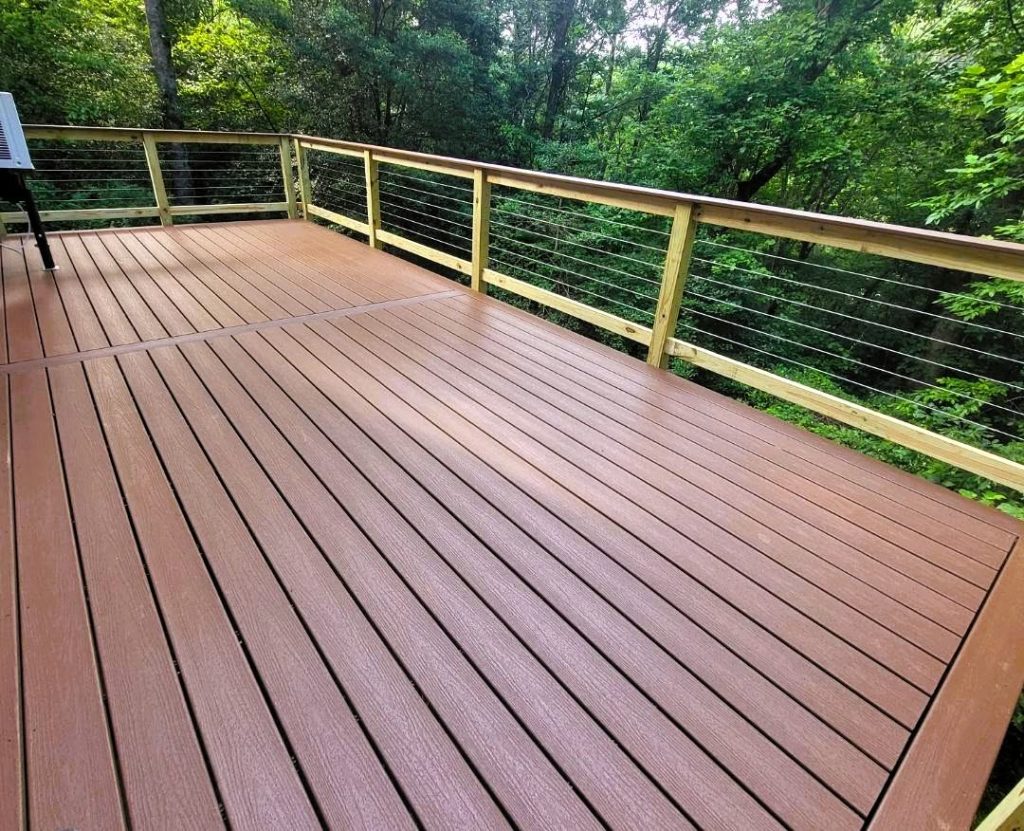 Red-brown composite deck with picture-frame border and pressure-treated cable railing facing dense green woods