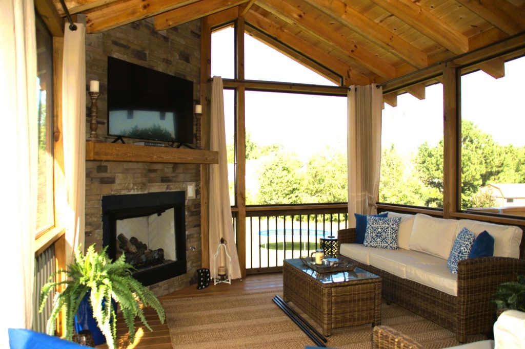 Screened porch interior with a stone fireplace, mounted television, cathedral pine ceiling and wicker seating overlooking a pool