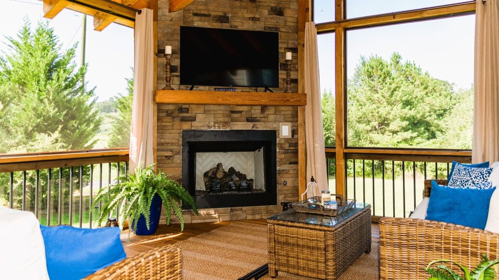 Screened porch built around a floor-to-ceiling stone fireplace with mounted television, timber mantel and wicker seating
