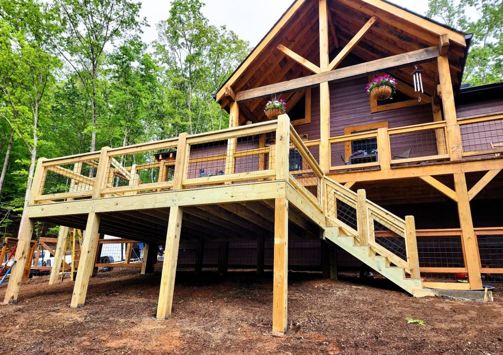 Large elevated pressure-treated deck with wire mesh railing wrapping a timber-frame cabin with a heavy gable truss