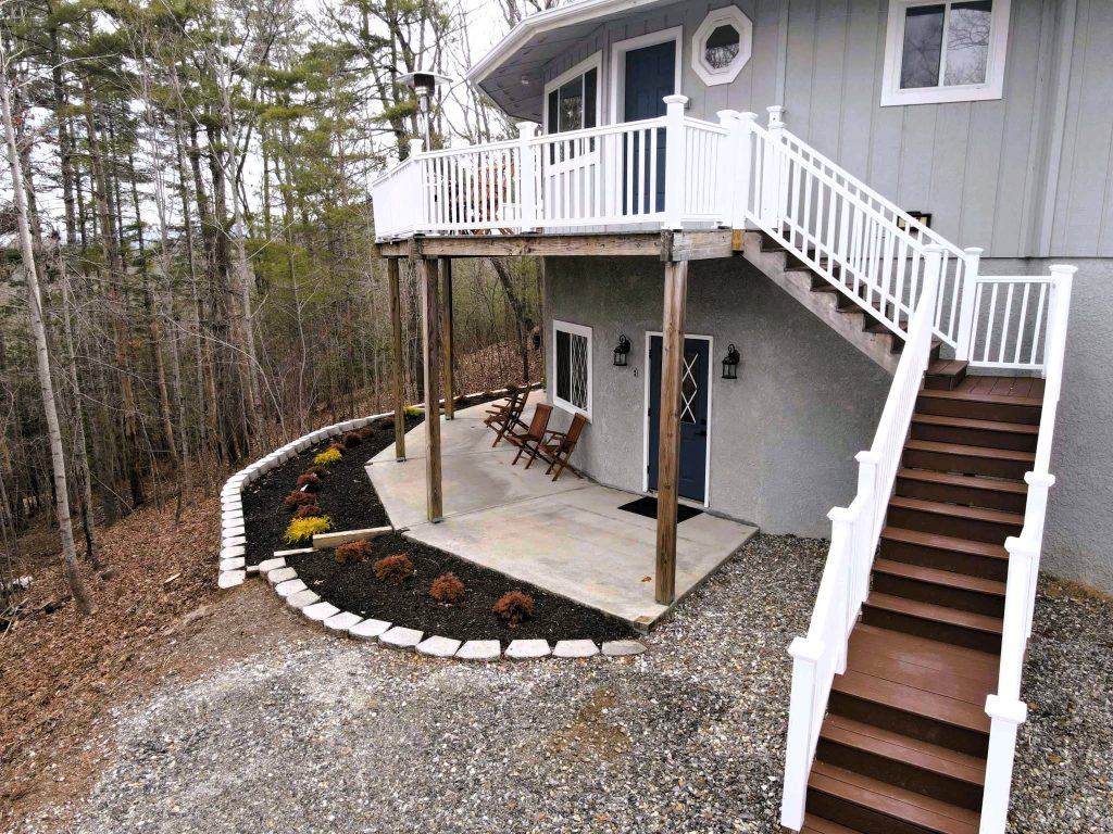 Elevated deck with white vinyl railing above a concrete patio, brown composite stairs descending past stone-edged planting beds